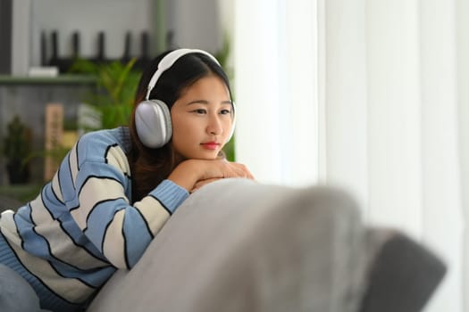 Thoughtful young woman listening to calm music in headphone and looking away while relaxing on couch.