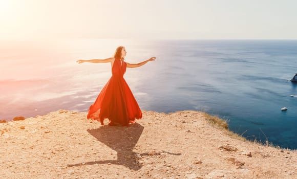 Side view a Young beautiful sensual woman in a red long dress posing on a rock high above the sea during sunrise. Girl on the nature on blue sky background. Fashion photo.