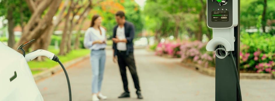 Focused EV car recharging battery on blurred background of lovey couple during autumnal road trip vacation by electric vehicle recharging battery. Eco friendly travel during autumn. Panorama Exalt