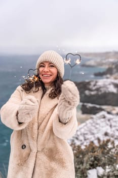 Outdoor winter portrait of happy smiling woman, light faux fur coat holding heart sparkler, posing against sea and snow background.