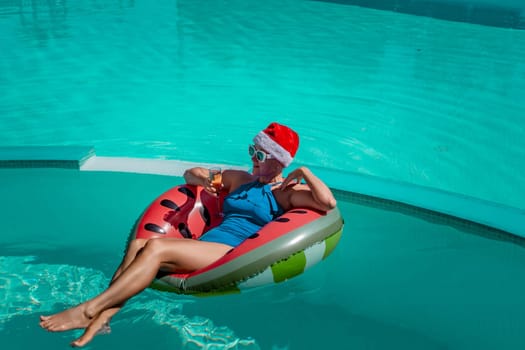 A happy woman in a blue bikini, a red and white Santa hat and sunglasses poses in the pool in an inflatable circle with a watermelon pattern, holding a glass of champagne in her hands. Christmas holidays concept