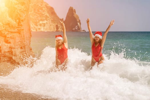 Women Santa hats ocean play. Seaside, beach daytime, enjoying beach fun. Two women in red swimsuits and Santa hats are enjoying themselves in the ocean waves and raising their hands up