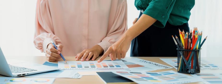 Cropped image of two female young beautiful graphic designer choose the color from color palate by using laptop on table with work tools and equipments scatter around at modern office. Variegated.