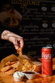 Ivano-Frankivsk, Ukraine March 26, 2023: Female hand dips chicken nuggets in sauce, chicken nuggets, french fries and coca cola on the table.