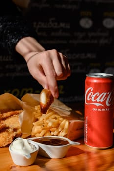 Ivano-Frankivsk, Ukraine March 26, 2023: Female hand dips chicken nuggets in sauce, chicken nuggets, french fries and coca cola on the table.