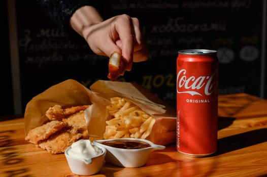 Ivano-Frankivsk, Ukraine March 26, 2023: Female hand dips chicken nuggets in sauce, chicken nuggets, french fries and coca cola on the table.