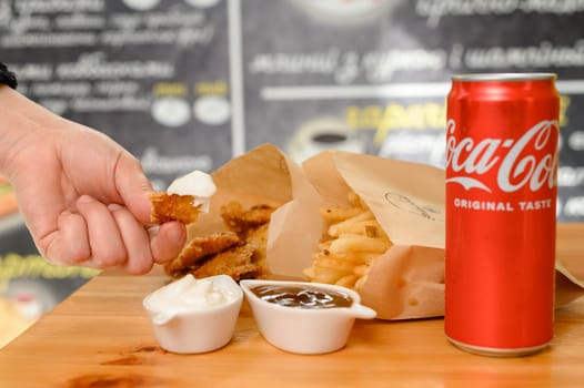 Ivano-Frankivsk, Ukraine March 26, 2023: Female hand dips chicken nuggets in sauce, chicken nuggets, french fries and coca cola on the table.