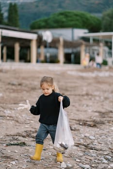 Little girl in gloves with a bag walks along the seashore collecting garbage. High quality photo