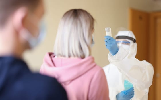 Person wearing protective medical suit and mask measures temperature with thermometer to visitors. Temperature control for coronavirus infection concept