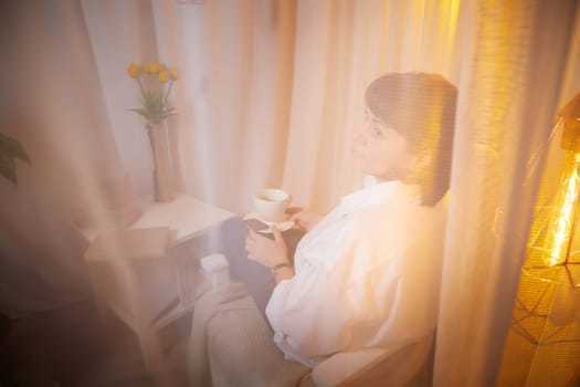 Nice girl with a mug of tea or coffee in cozy room. A middle-aged adult woman relaxing in the evening in the living room or bedroom in the evening