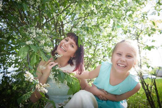 Happy mother and daughter enjoying rest, playing and fun on nature on a green lawn and with blooming apple tree in the background. Woman and girl resting outdoors in summer and spring day