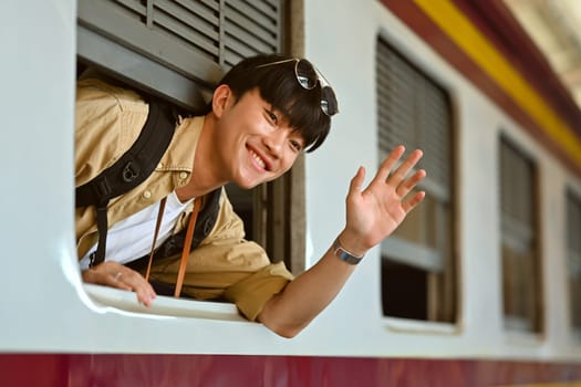 Happy Asian male backpacker waving from the train window. Transportation, people and traveling concept