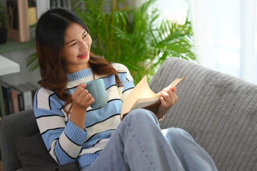 Cheerful young woman reading book and drinking hot tea on couch at home.