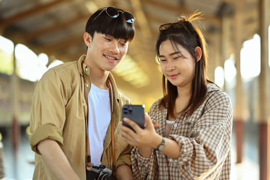 Cheerful Asian couple tourist checking timetable train schedule for trip on mobile phone.