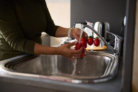 Close-up of a woman housewife washing ripe organic tomato cherry under a tap with running water, in the home kitchen