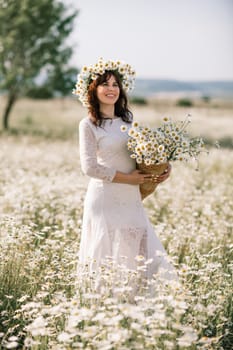 Happy woman in a field of daisies with a wreath of wildflowers on her head. woman in a white dress in a field of white flowers. Charming woman with a bouquet of daisies, tender summer photo.