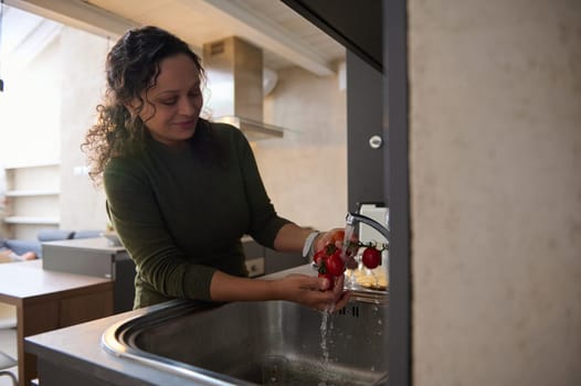 Beautiful curly brunette young woman, pleasant housewife, standing at sink and washing fresh tomatoes under running water, preparing healthy meal in the home kitchen. People. Lifestyle. Domestic life