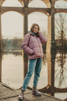 A young woman standing at the shore looking at the river in autumn sunny day. Street view, copy space for text, travel photo. Happy tourist woman on the bank of the river in autumn in warm clothes. Tourists enjoy their vacation, winter season. Romantic look and travel concept. A joyful mood in a Caucasian girl. Winter Wonderland: Enchanting Girl by the Riverside in Autumn.