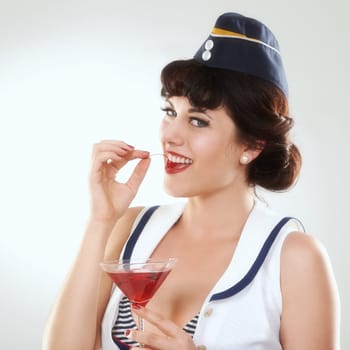 Portrait, stewardess and happy woman with alcohol to drink in studio isolated on white background. Face, martini cocktail glass and air hostess eating cherry, travel or vintage pin up girl on journey.