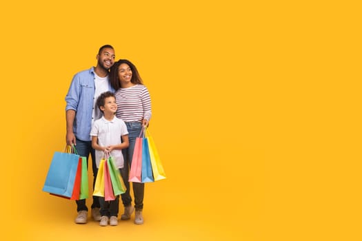 Cheerful black family of three with colorful shopping bags in hands looking aside at copy space, happy african american mother, father and son enjoying seasonal sales, posing on yellow background