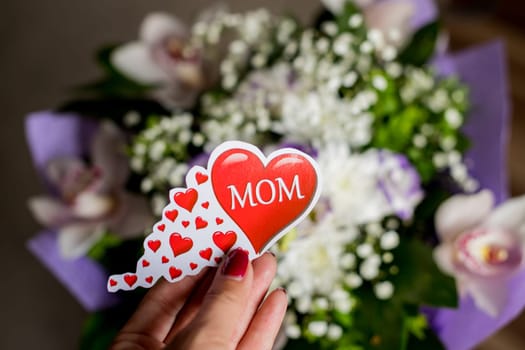 Bouquet of orchids, Lisianthus,Gypsophila.Female hand is holding a big red heart with text mom, Present with love.