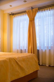 Luxury interior of bedroom with big window with white curtains. Comfortable bed with sheets and yellow pillows in modern style.
