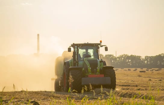 Combine harvester pressing straw from field into bales driving field on sunny summer evening. Field with bales of pressed wheat. Lots of dust on field. Agricultural agro-industrial harvesting works