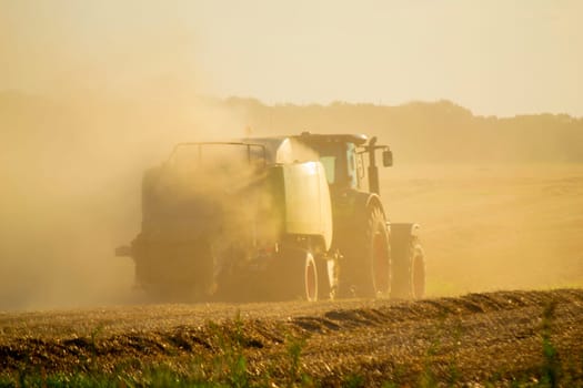 Combine harvester pressing straw from field into bales driving field on sunny summer evening. Field with bales of pressed wheat. Lots of dust on field. Agricultural agro-industrial harvesting works