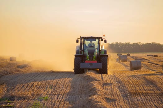 Combine harvester pressing straw from field into bales driving field on sunny summer evening. Field with bales of pressed wheat. Lots of dust on field. Agricultural agro-industrial harvesting works