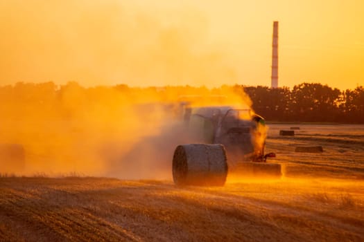 Combine harvester pressing straw from field into bales driving field on sunny summer evening. Field with bales of pressed wheat. Lots of dust on field. Agricultural agro-industrial harvesting works