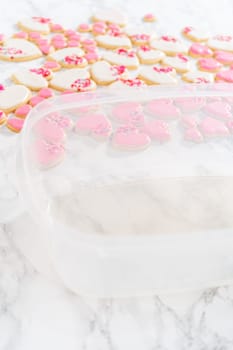 Storing heart-shaped sugar cookies with pink and white royal icing in a large plastic container.