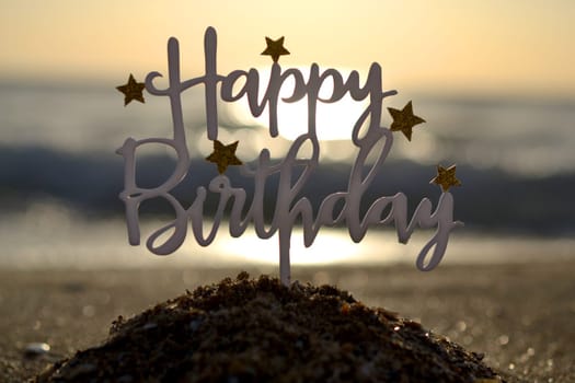 Stick in shape of letters Happy Birthday with stars at dawn and sunset on background of sea waves on sand seashore. Word Happy birthday in sandy beach on sunny summer day. Concept Happy Birthday