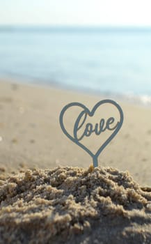 Plastic stick in shape of heart and word Love in sand on beach seashore on sunny summer day close-up. Figures in shape of heart word Love on background of sea waves. Love relationship romance concept