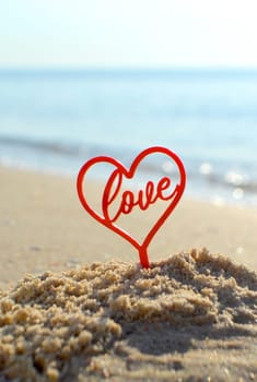 Plastic stick in shape of red heart and word Love in sand on beach seashore on sunny summer day close-up. Figures in shape of heart word Love on background sea waves. Love relationship romance concept