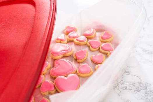 Storing heart-shaped sugar cookies with pink and white royal icing in a large plastic container.