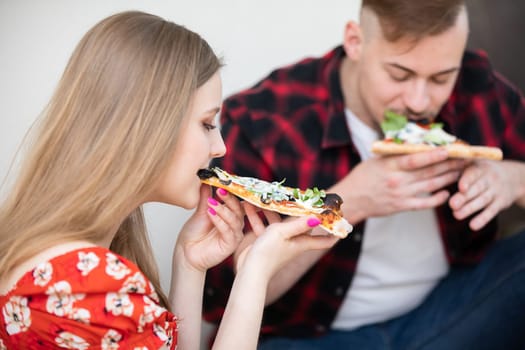 A close-up of a young woman's profile in the foreground. She has her hair down. The woman is biting into a slice of pizza. A man can be seen in the background, his figure is blurred. He is also eating a pizza.