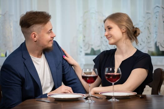 The girl strokes her partner's shoulder. The couple is sitting close together at a table. Two glasses of red wine stand on the table. The couple is elegantly dressed.