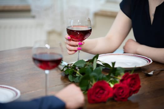 Two glasses of red wine stand on the table next to a bouquet of roses. The woman holds one of the glasses. She has her nails painted with pink nail polish.