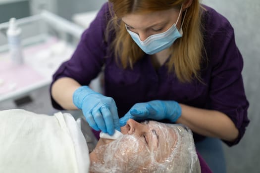 The cosmetologist works with blue disposable gloves. A woman lies on a cosmetic bed in an aesthetic clinic. Her face is partially wrapped in a special film.