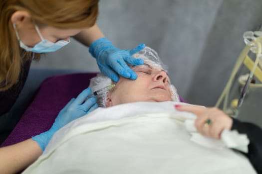 Close-up of a silhouette of a mature woman lying on a bed under a cover. The woman is wearing a disposable cap on her head. A grimace of pain is visible on her face. A nurse stands next to her and performs an injection.