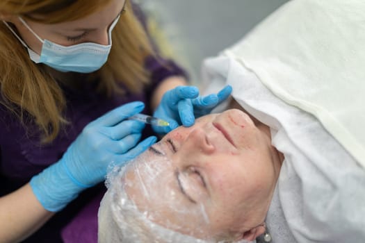 View from above. A mature woman lies on a bed covered with a white sheet. She is wearing a protective cap on her head. Next to her sits a nurse wearing a surgical mask and administering an injection.