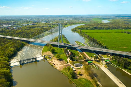 Massive pylon bridge spanning Oder river flowing by Wroclaw. Redzinski Bridge and lush greenery around city of Poland on sunny day aerial view