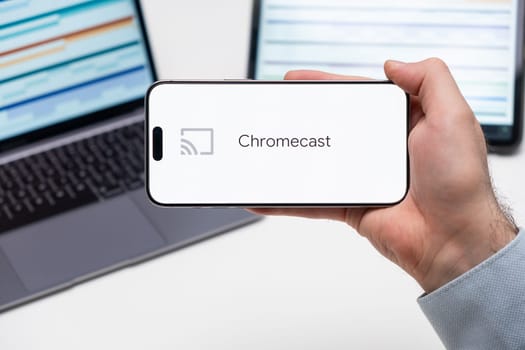 Chromecast logo of app on the screen of mobile phone held by man in front of the laptop and tablet, December 2023, Prague, Czech Republic