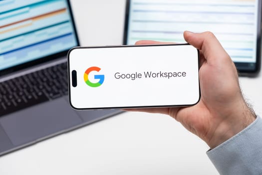 Google Workspace application logo on the screen of smart phone in mans hand, laptop and tablet are on the table in the background, December 2023, Prague, Czech Republic.
