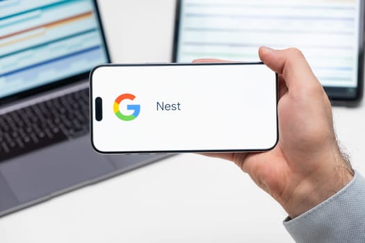 Nest application logo on the screen of smart phone in mans hand, laptop and tablet are on the table in the background, December 2023, Prague, Czech Republic.
