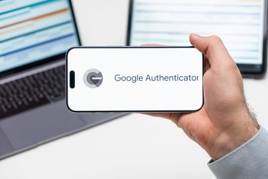 Google Authenticator logo of app on the screen of mobile phone held by man in front of the laptop and tablet, December 2023, Prague, Czech Republic