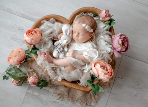Newborn Baby Girl Sleeps In Heart-Shaped Wooden Bowl Among Flowers During Studio Photoshoot