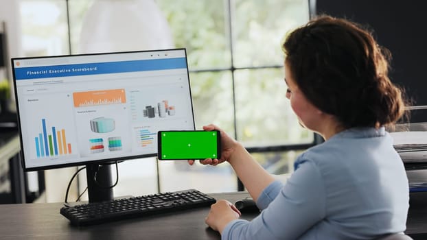 Businesswoman looks at greenscreen while she holds mobile device, isolated chromakey display shown at office desk. Employee checking blank copyspace software on phone. Handheld shot.