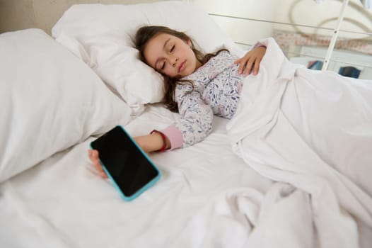 Caucasian child girl in pajamas, falling asleep on her bed with a mobile phone in her hand. Smartphone with black blank mockup digital touch screen. Digital gadget and online video games addiction