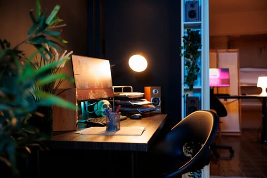 Work desk with computer and house plants in empty home studio interior with warm lighting. Apartment illuminated by neon lights with 3D rendered animations running on powerful PC monitors